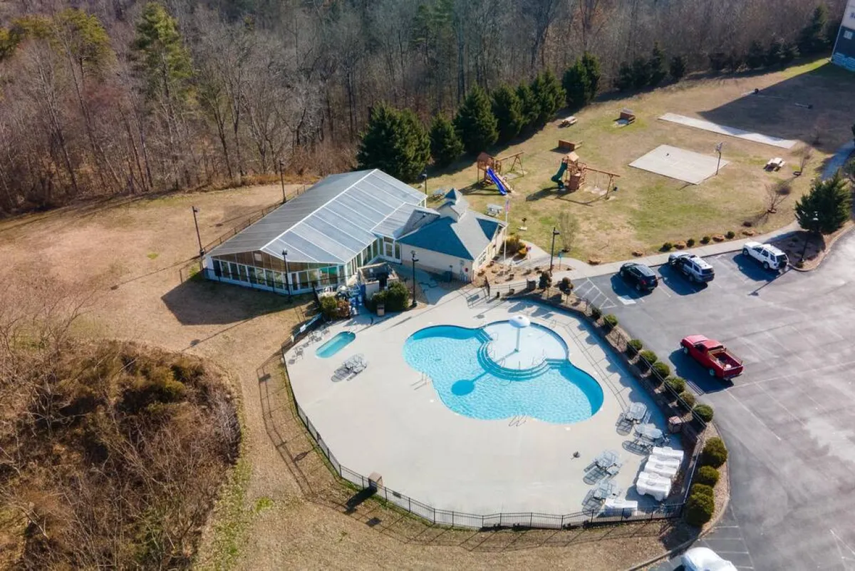 An outdoor scene featuring a large swimming pool, a visible playground, and a community center, with children playing and a parking lot in the background. The image captures a lively, family-friendly atmosphere on the resort grounds.