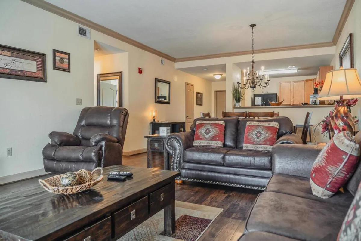 A spacious living room featuring leather sofas arranged around a wood coffee table, adorned with decorative pillows and illuminated by a chandelier. The open layout and inviting arrangement encourage relaxation and social interaction, ideal for family gatherings or movie nights.