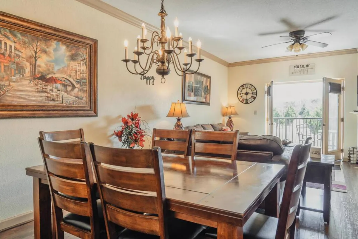 A dining table set under a chandelier, adjacent to a cozy sofa and with a view of the balcony. The setup encourages group gatherings, family dinners, and socializing, with natural light and outside visibility.