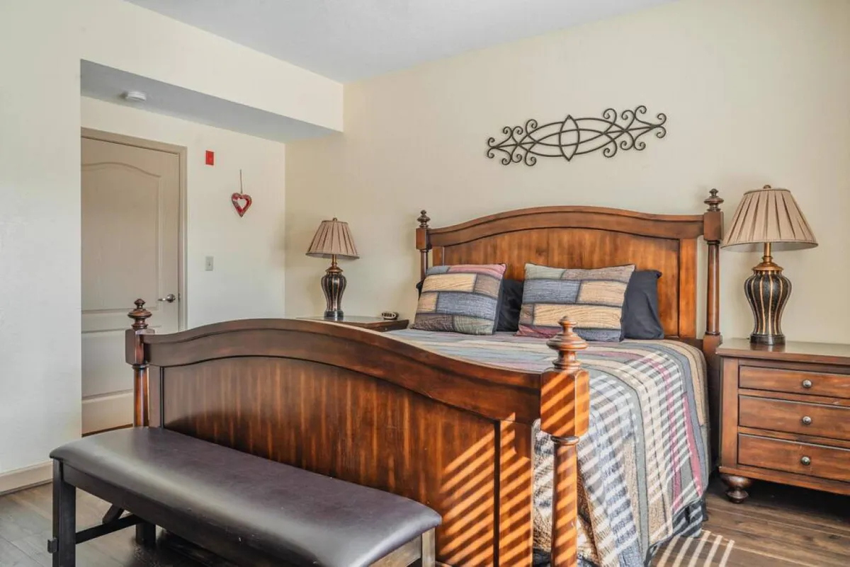 The image shows a cozy bedroom interior featuring a sturdy wooden bed frame with inviting striped bedding, flanked by warm bedside lamps and a nightstand. Soft natural light filters into the room, creating a welcoming and comfortable atmosphere ideal for relaxation.