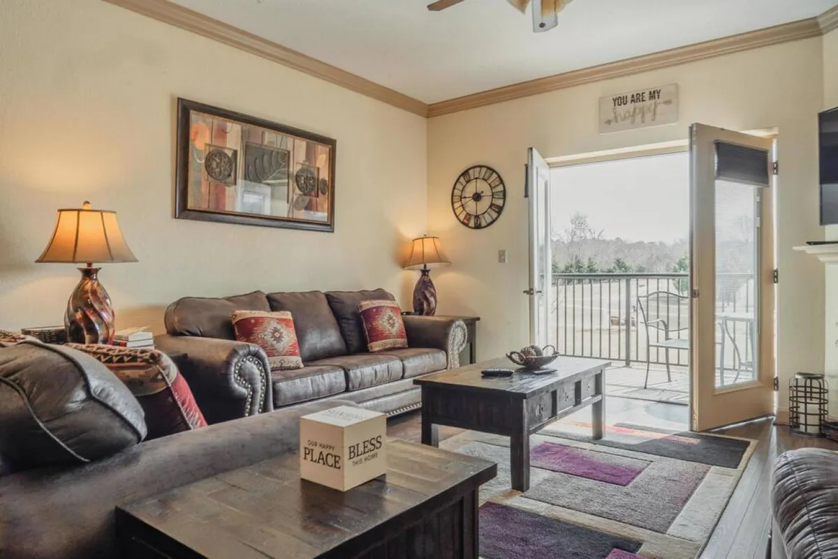 A cozy living area featuring a leather sofa, coffee table, decorative lamps, wall art, and a door leading out to a balcony. The space is filled with natural light and offers a glimpse of outdoor views, suggesting a connection to the surrounding area.