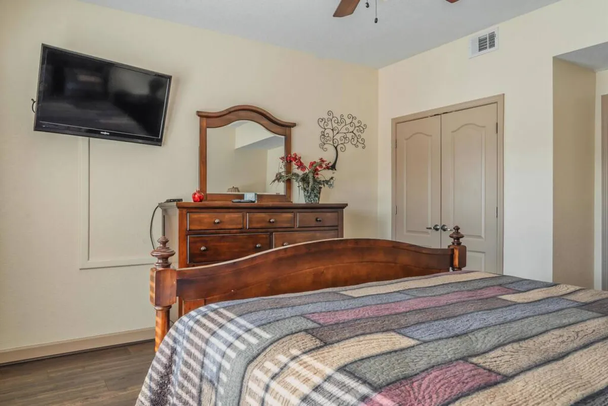 A cozy bedroom with a wooden bed frame, dresser with mirror, wall-mounted TV, decorative wall art, and a flower arrangement. Natural light streams into the room, enhancing the sense of comfort and calm for a restful night's sleep or relaxed morning.