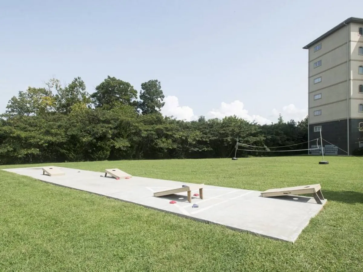 A vibrant outdoor area featuring cornhole boards set up on green grass, surrounded by trees and a building in the distance. The scene conveys energy and playfulness, inviting families to enjoy lawn games and open space activities together.