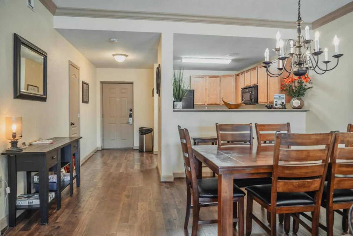 A wide-angle view of a dining area with a table and chandelier, adjacent to a kitchen and the condo’s entrance door. The setup suggests easy movement to and from the condo, ideal for guests coming and going on daily adventures.
