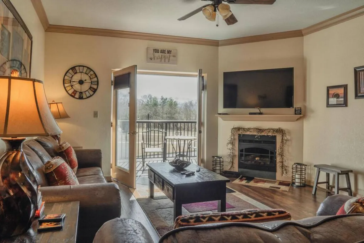 A cozy living space with a comfortable sofa, coffee table, inviting fireplace, open doors to a balcony, and a wall-mounted flat-screen TV. The space is bathed in natural light, blending indoor comfort with access to outdoor views.